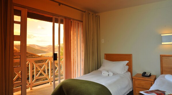 A hotel room with a bed, a nightstand, a telephone, and a view of mountains through the window. - Mohale Lodge (Likalaneng)