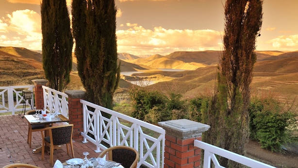 An outdoor dining area with tables and chairs, overlooking a valley with hills and a lake.