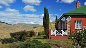 A red brick house with satellite dishes, surrounded by a lush garden and hills in the background.