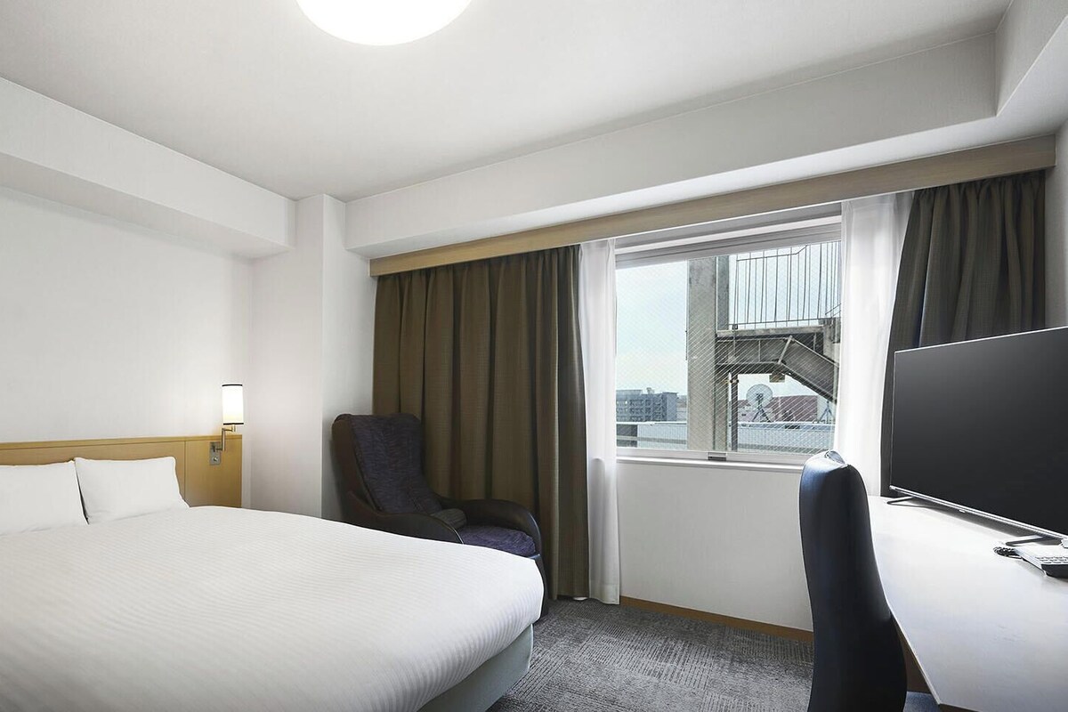 A hotel room with a bed, a desk with a TV, a chair, and a window with curtains.