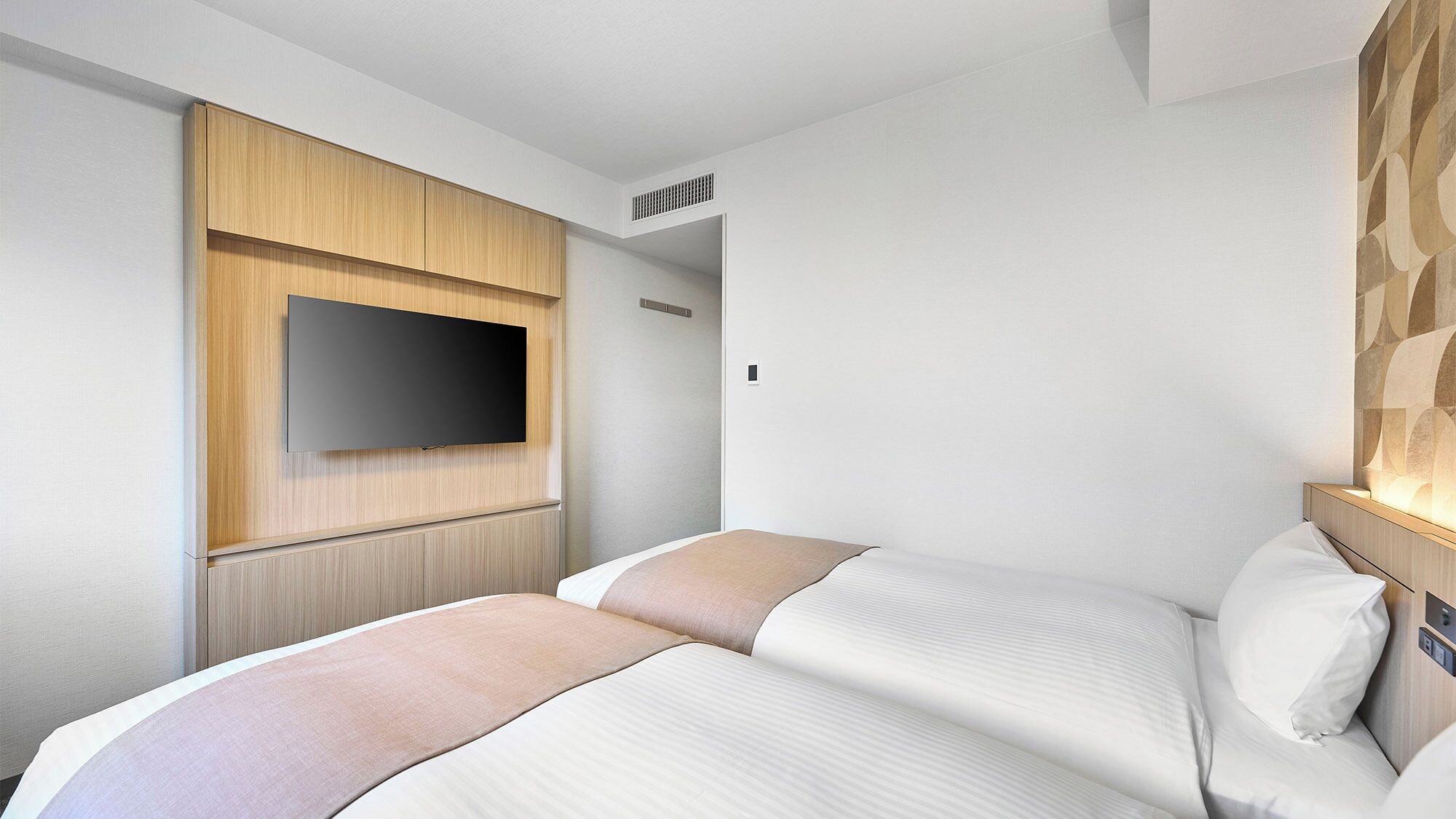 A hotel room with a bed, a flat-screen TV mounted on a wooden panel, and a wall with a geometric pattern.