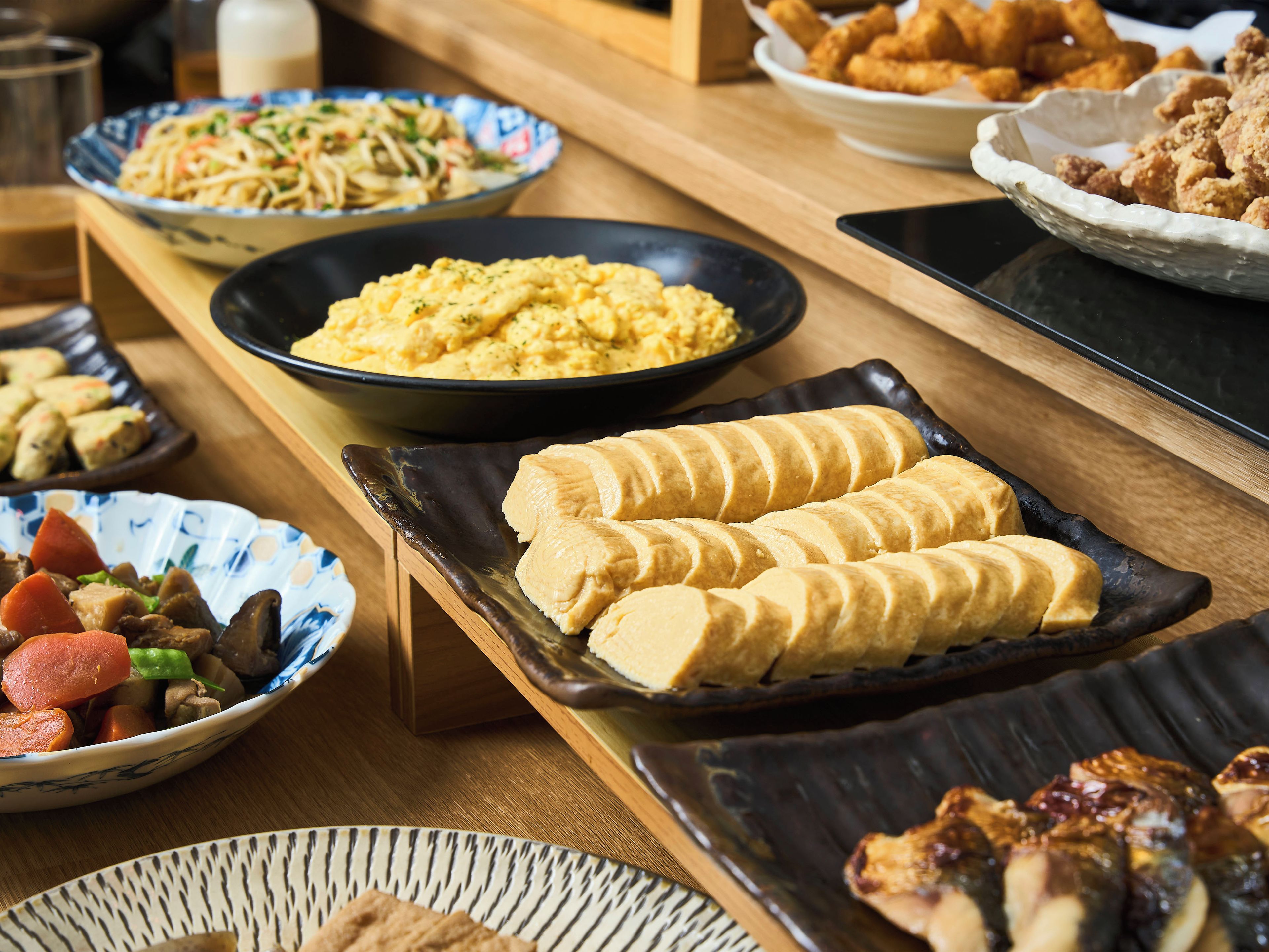 A variety of dishes on a wooden table, including a bowl of noodles, a plate of scrambled eggs, and several dishes of fried food.