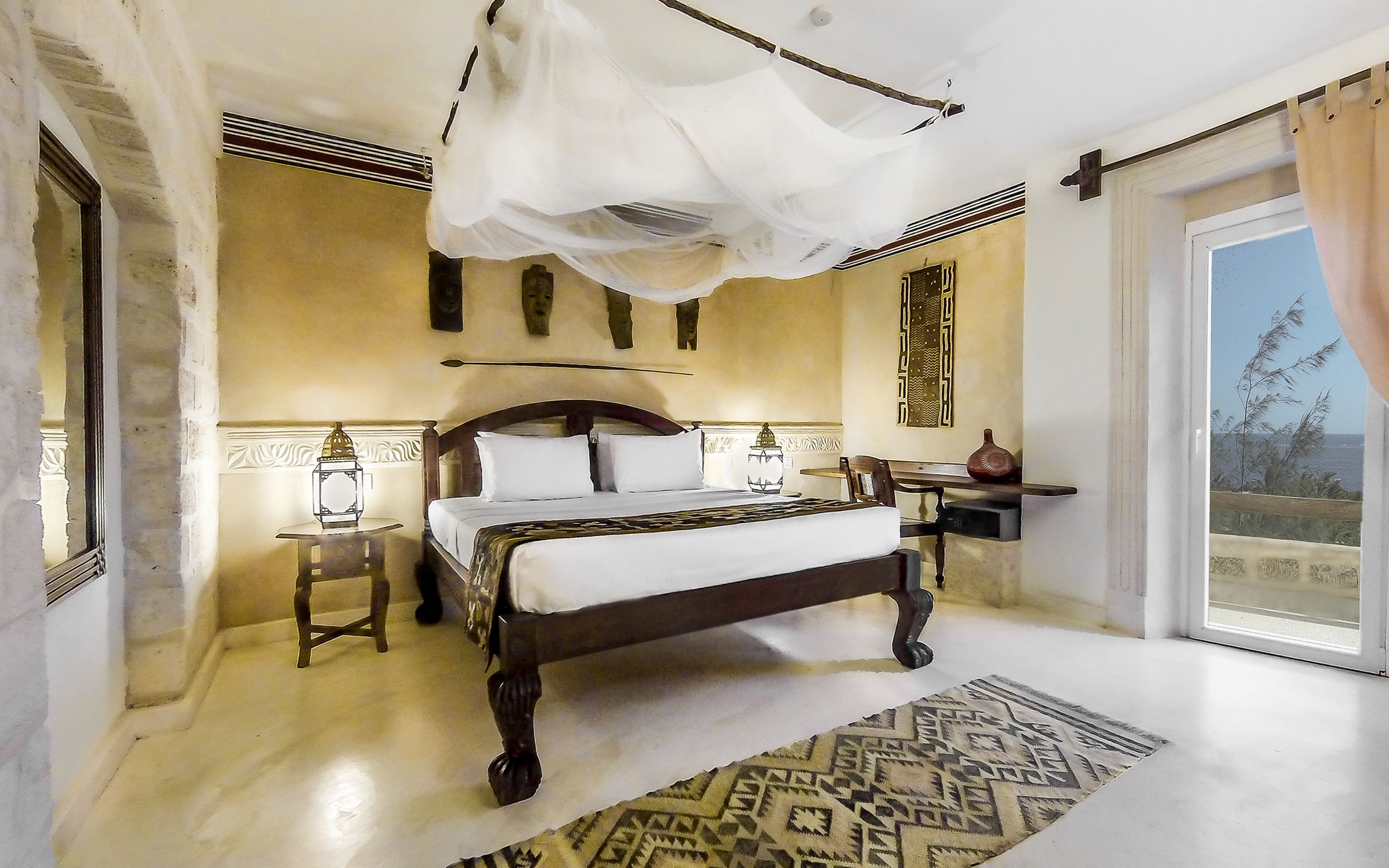 A bedroom with a large bed, a nightstand, a lamp, and a view of the sea.