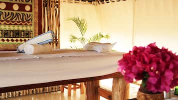 Relax in a cozy room with a wooden bed, a table, and a vase with flowers, set amidst a thatched roof and patterned walls.