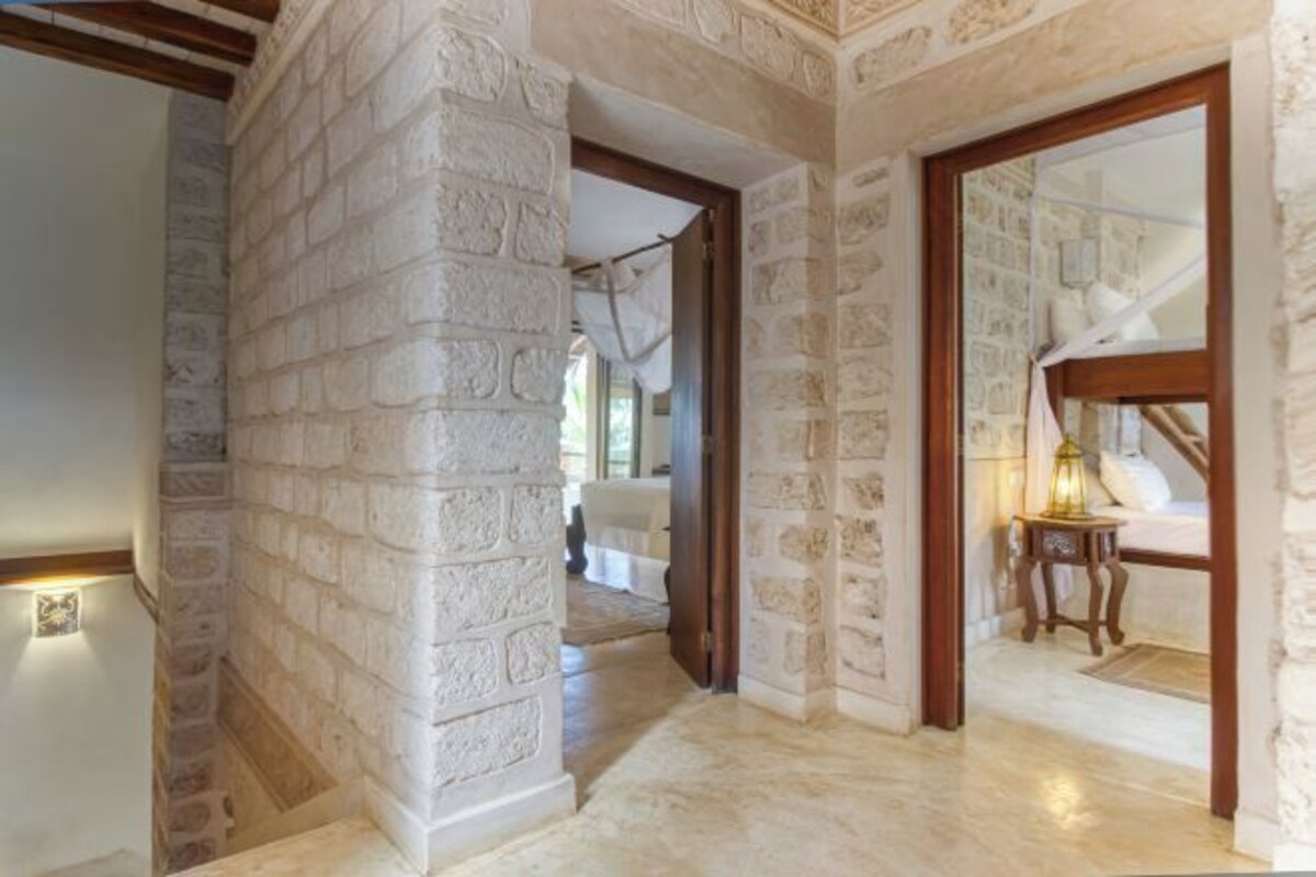A hallway with stone walls and wooden beams, leading to a bedroom with a bed and a nightstand.