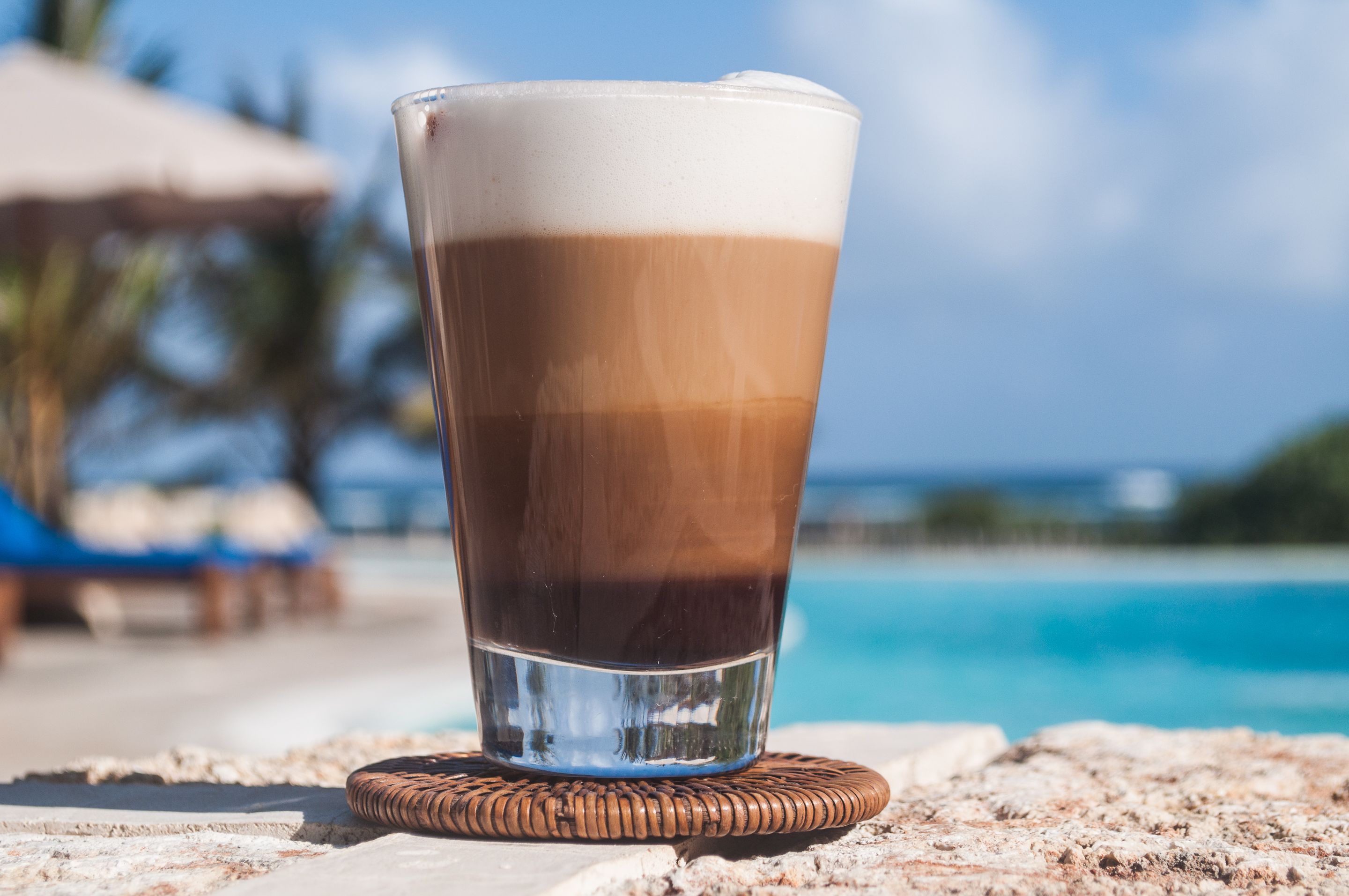 Un verre de café glacé, posé sur un sous-verre en fibres tressées, près d’une piscine.