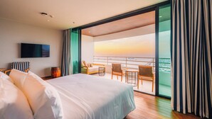 Een moderne hotelkamer met een groot bed, een flatscreen televisie, een balkon met uitzicht op de zonsondergang en een zithoek met stoelen en een tafel.