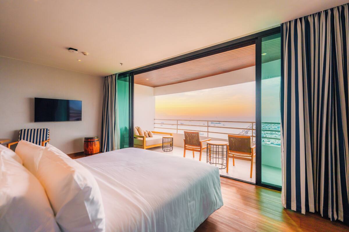 Een moderne hotelkamer met een groot bed, een flatscreen televisie, een balkon met uitzicht op de zonsondergang en een zithoek met stoelen en een tafel.