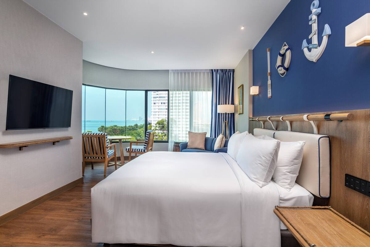 Een moderne hotelkamer met een groot bed, een flatscreen televisie, een zithoek met een tafel en stoelen, en een uitzicht op de skyline door grote ramen.
