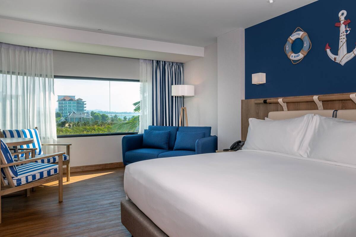 Een hotelkamer met een groot bed, een blauwe bank, een gestreepte fauteuil, een nachtkastje en uitzicht op een gebouw door het raam.
