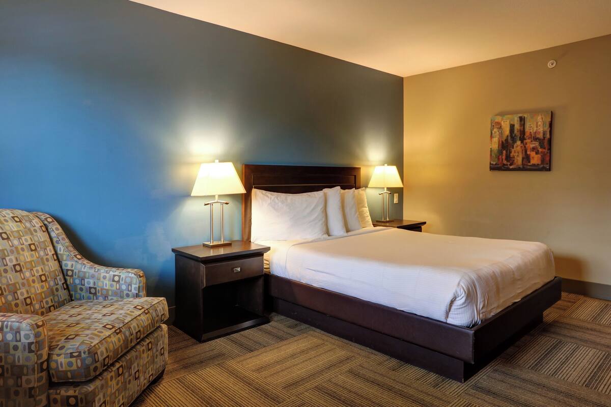 A hotel room with a bed, two bedside tables with lamps, a chair, and a painting on the wall.
