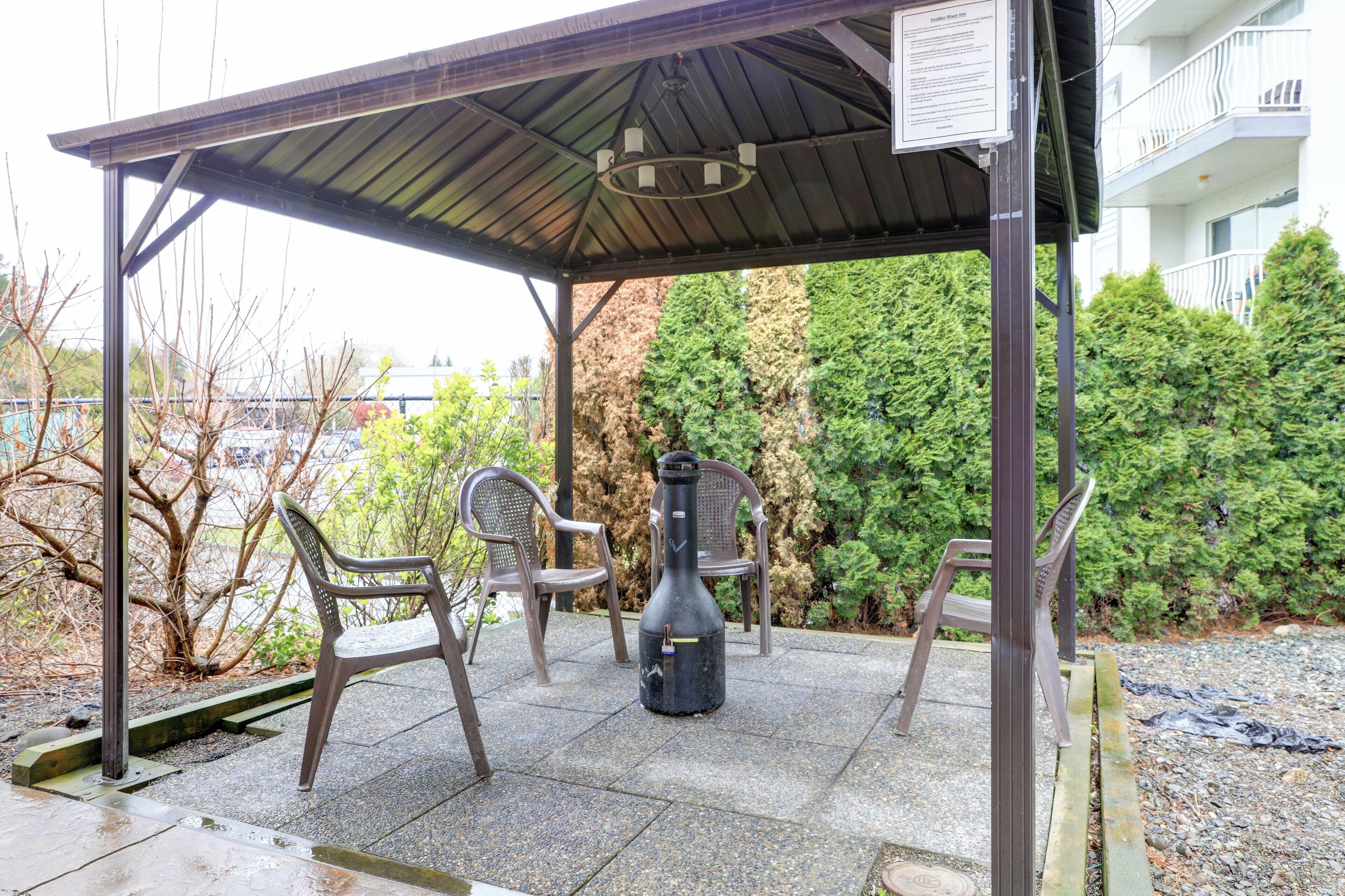 Un espacio de estar al aire libre techado con sillas y una guitarra, rodeado de vegetación y un edificio al fondo.