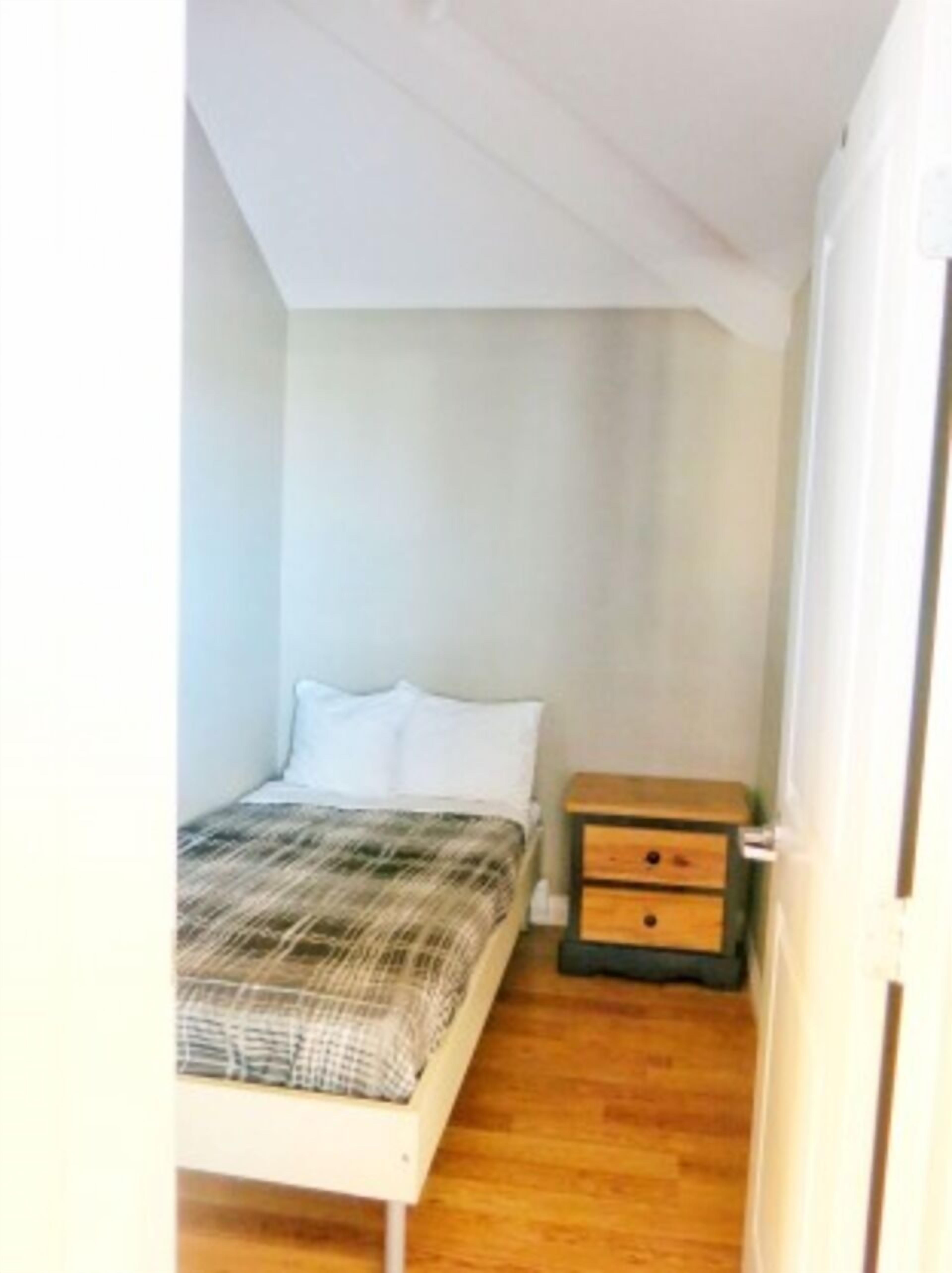 Habitació petita amb un llit individual, sòl de fusta, capçal de llit blanc i una tauleta de nit amb calaixos.