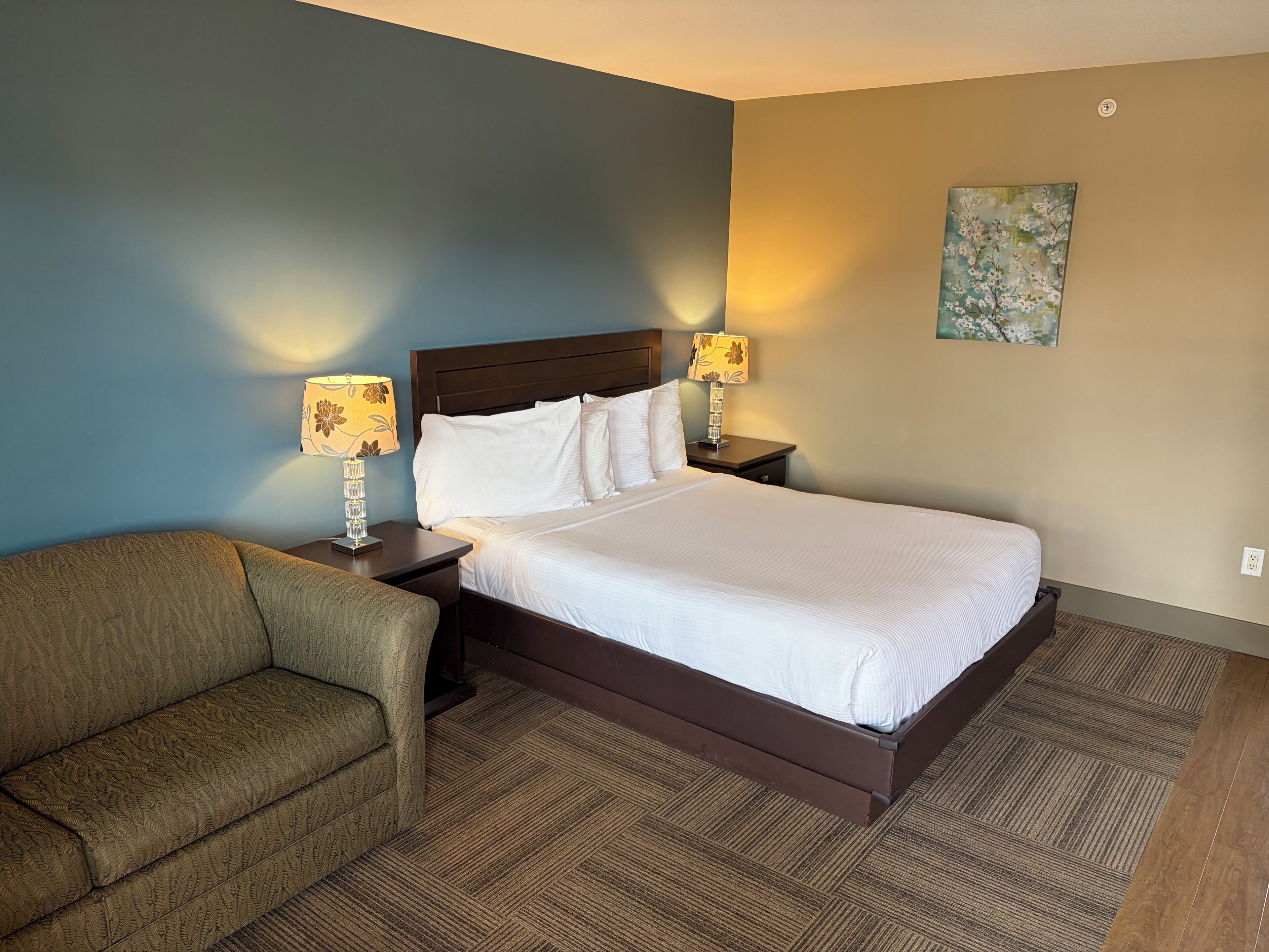 Una habitación de hotel con una cama, dos mesitas de noche con lámparas, una silla y un cuadro en la pared.