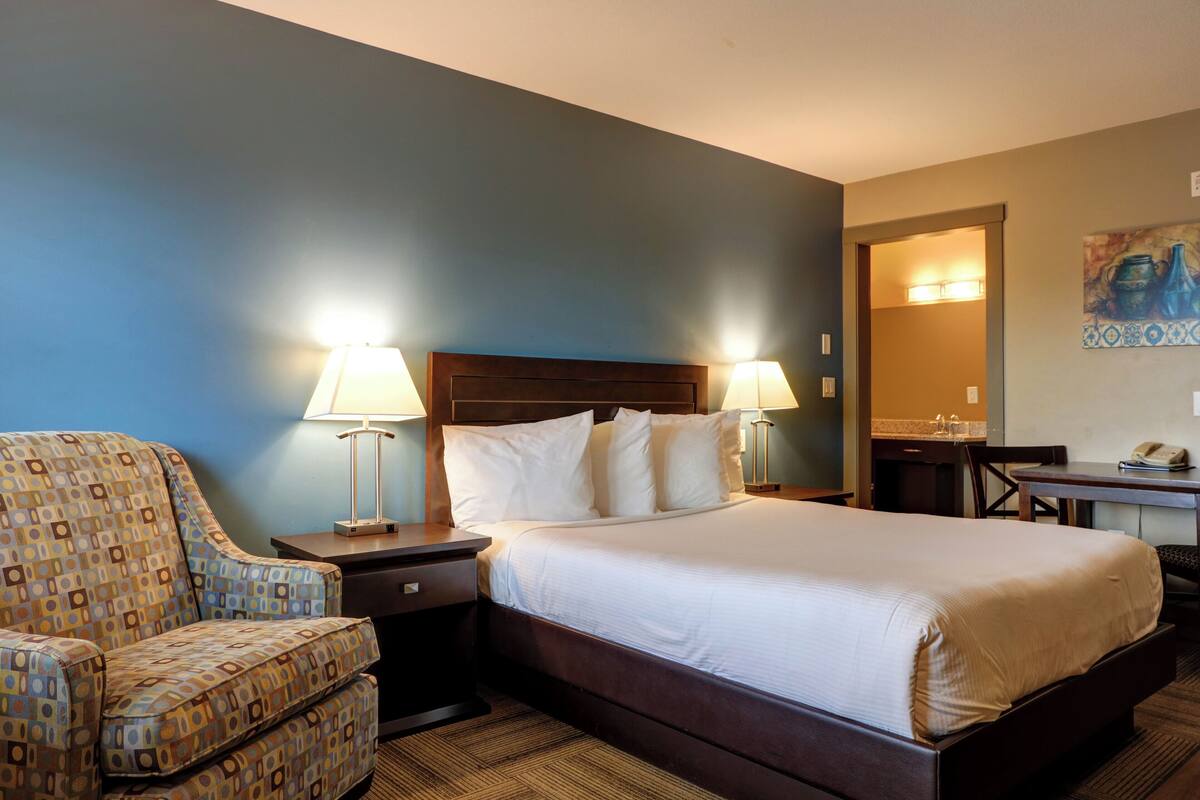 A hotel room with a bed, two bedside lamps, a patterned armchair, a desk, and a painting on the wall.