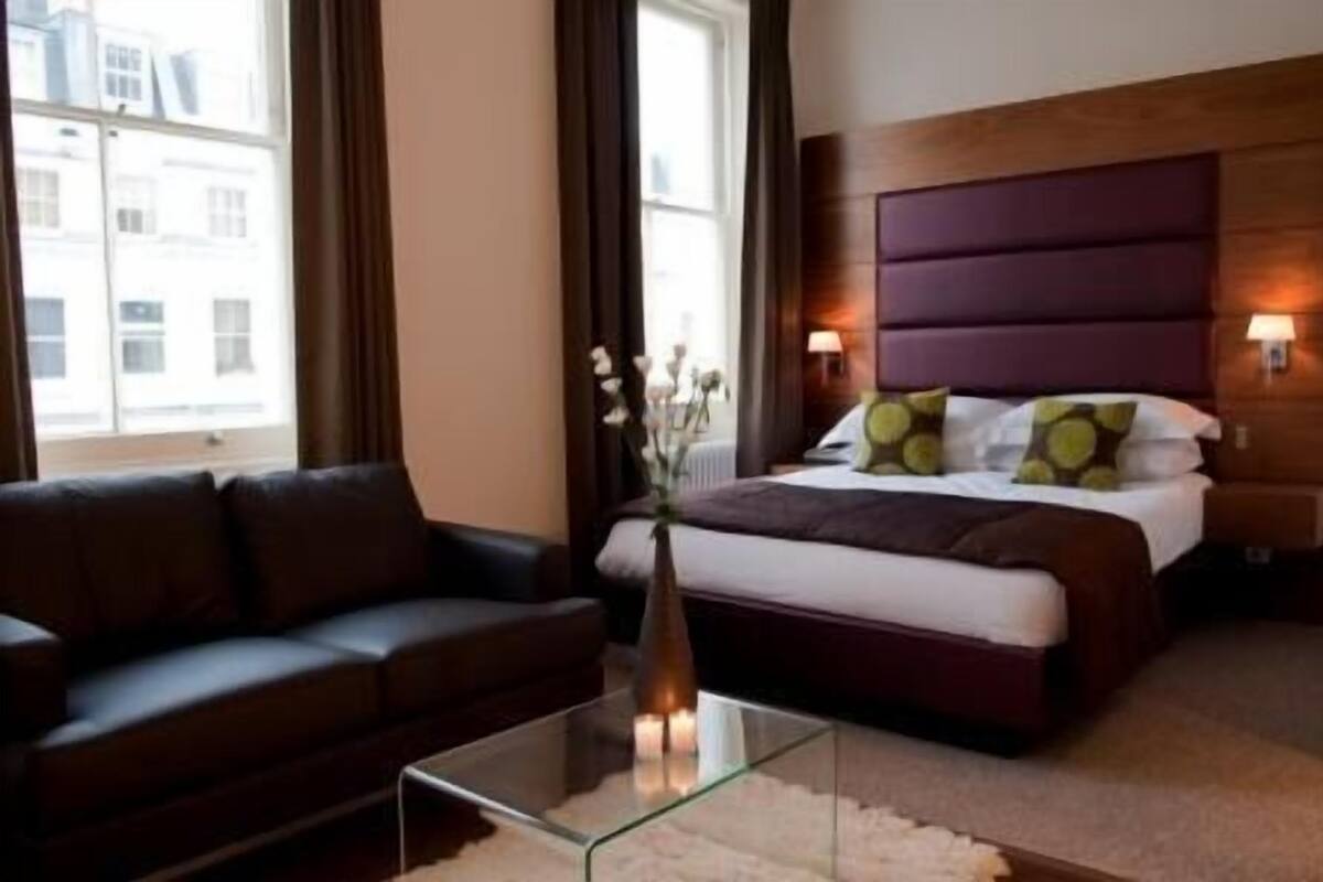 A hotel room with a bed, a sofa, a glass coffee table, and a vase with flowers.
