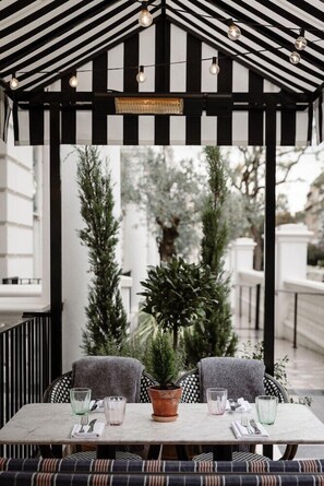An outdoor seating area with a table and chairs, a potted plant, and a pergola with hanging lights. - 10 Pembridge Gardens (London)