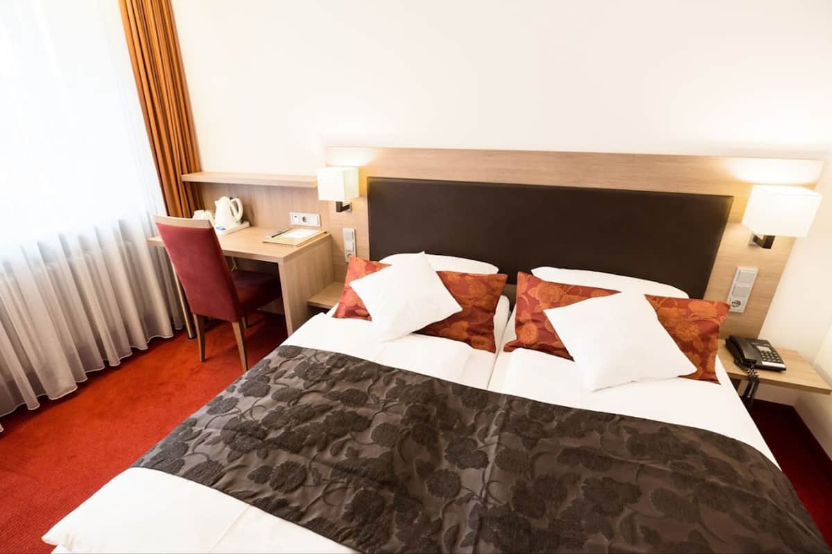 A hotel room with a bed, a desk, a chair, a telephone, and a lamp.