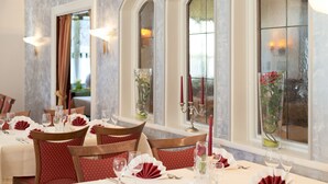 A dining area with tables set for a meal, featuring red and white tablecloths, candles, and glass vases with flowers.
