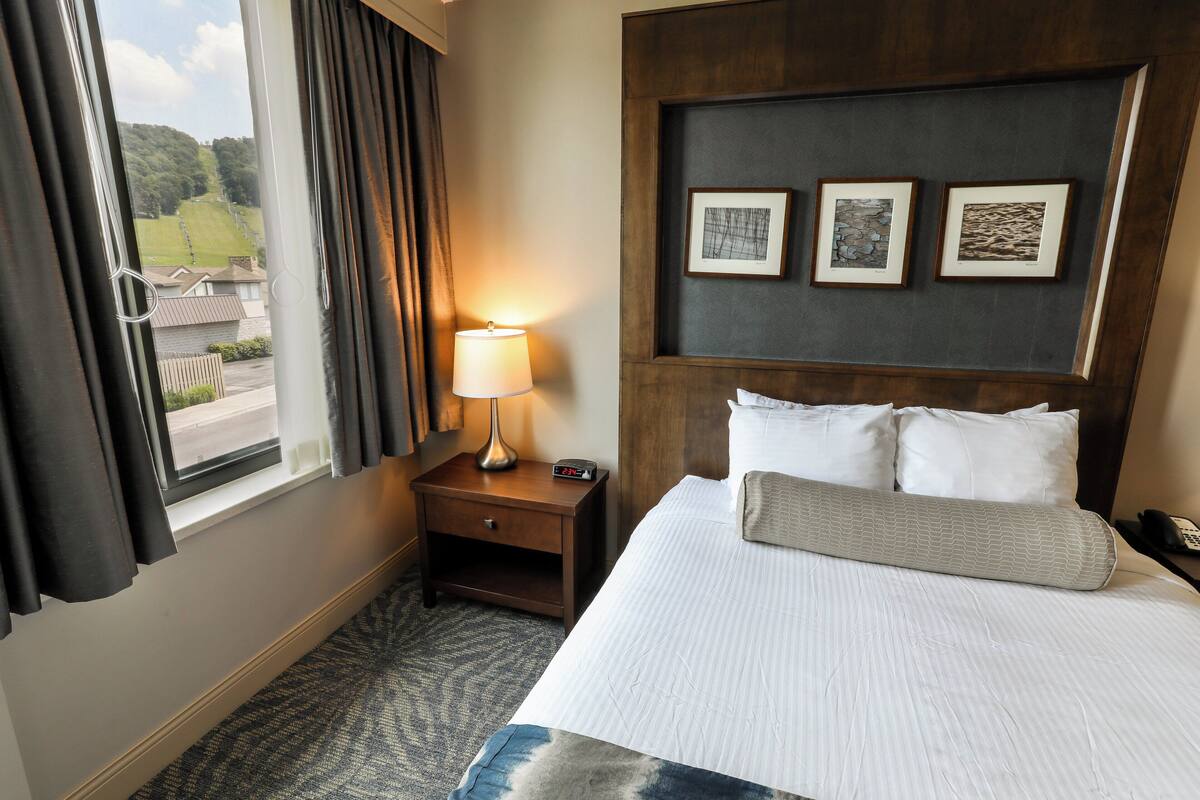 A hotel room with a bed, a nightstand with a lamp, a wall-mounted phone, and three framed pictures above the bed.