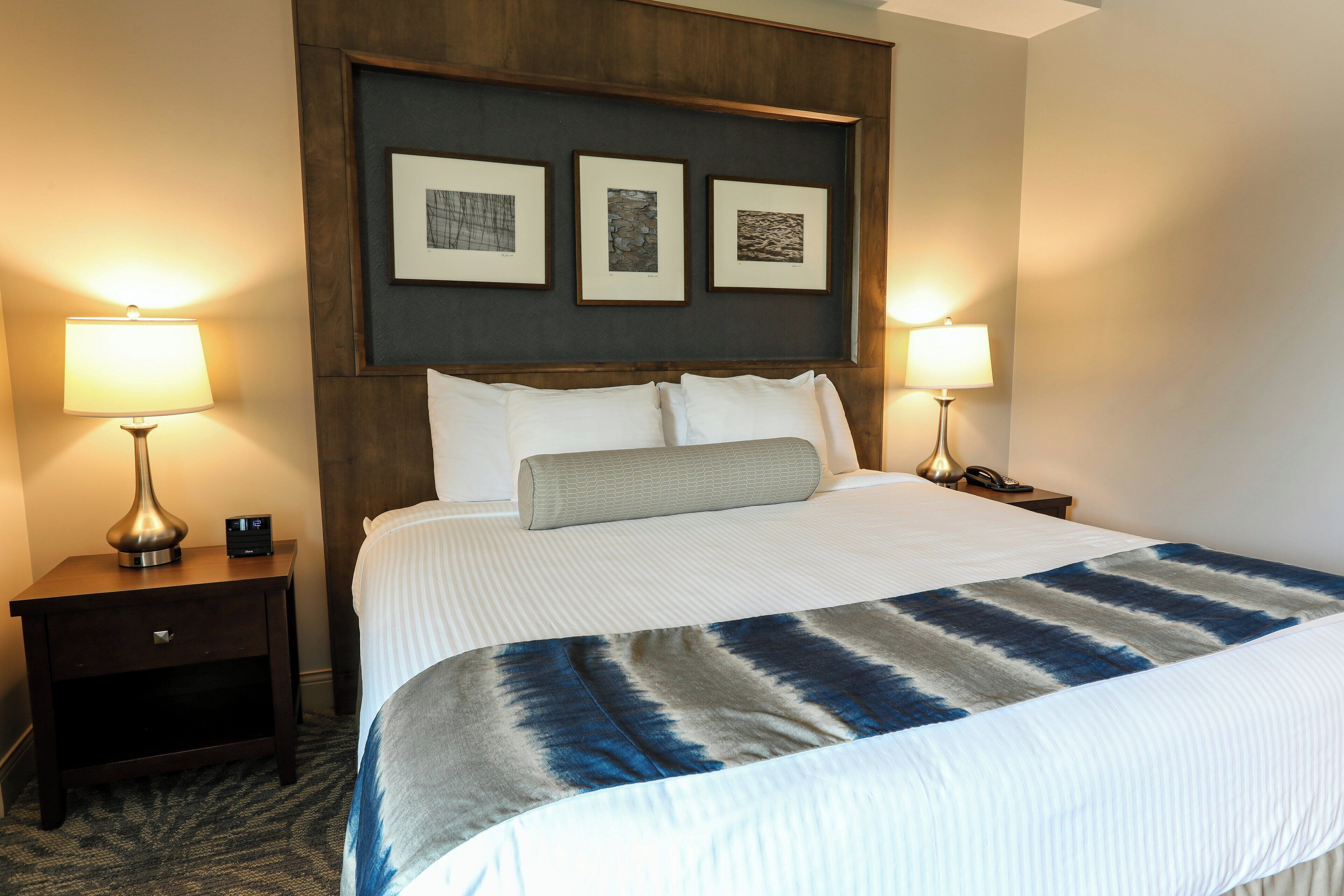 Habitación de hotel con una cama grande, dos mesitas de noche con lámparas, un cabecero con cuadros enmarcados y piso alfombrado.