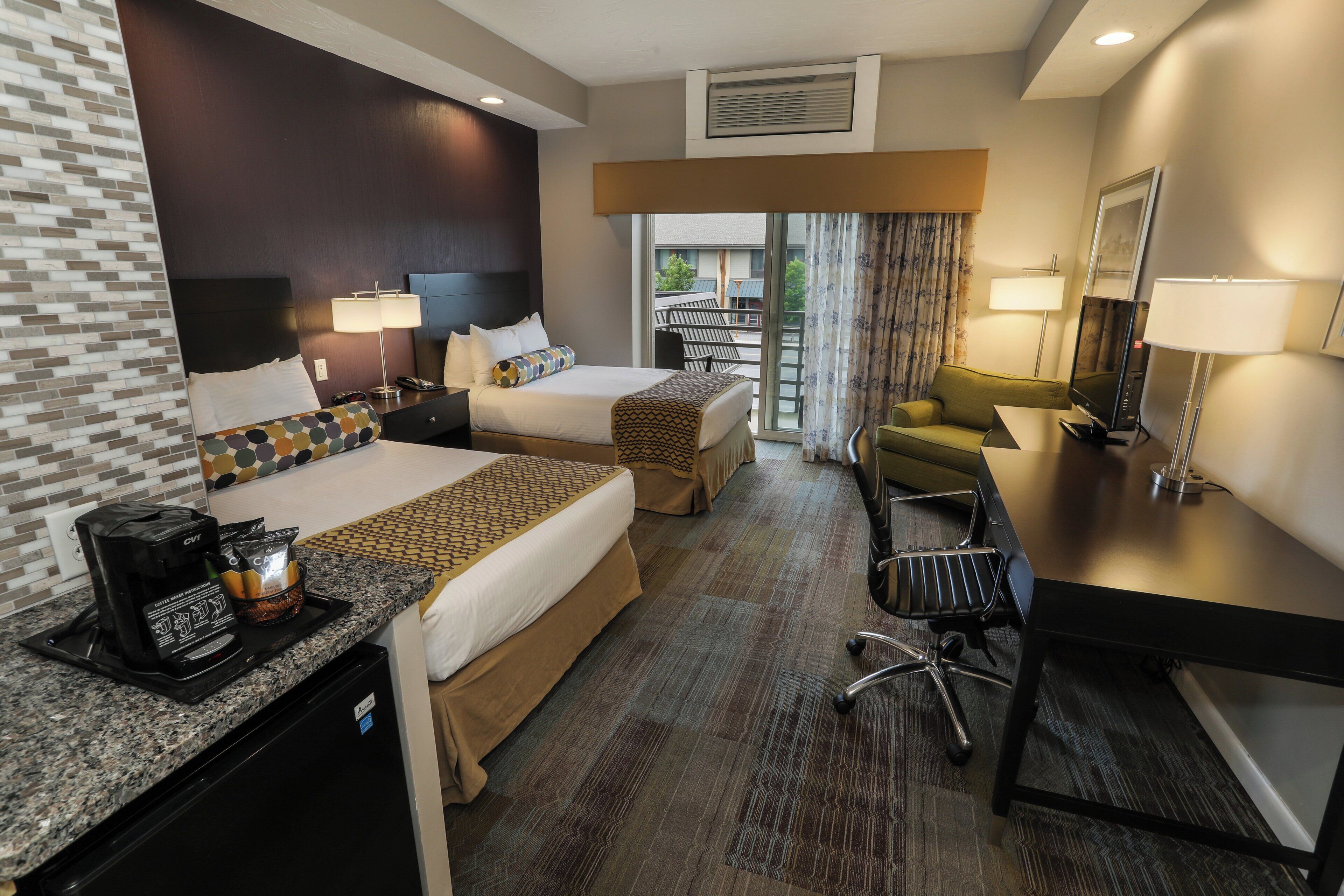 A hotel room with two beds, a desk with a coffee maker, a chair, and a view of the outdoors.