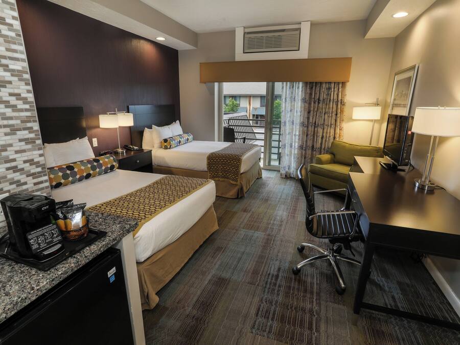 A hotel room with two beds, a desk with a coffee maker, a chair, and a view of the outdoors.
