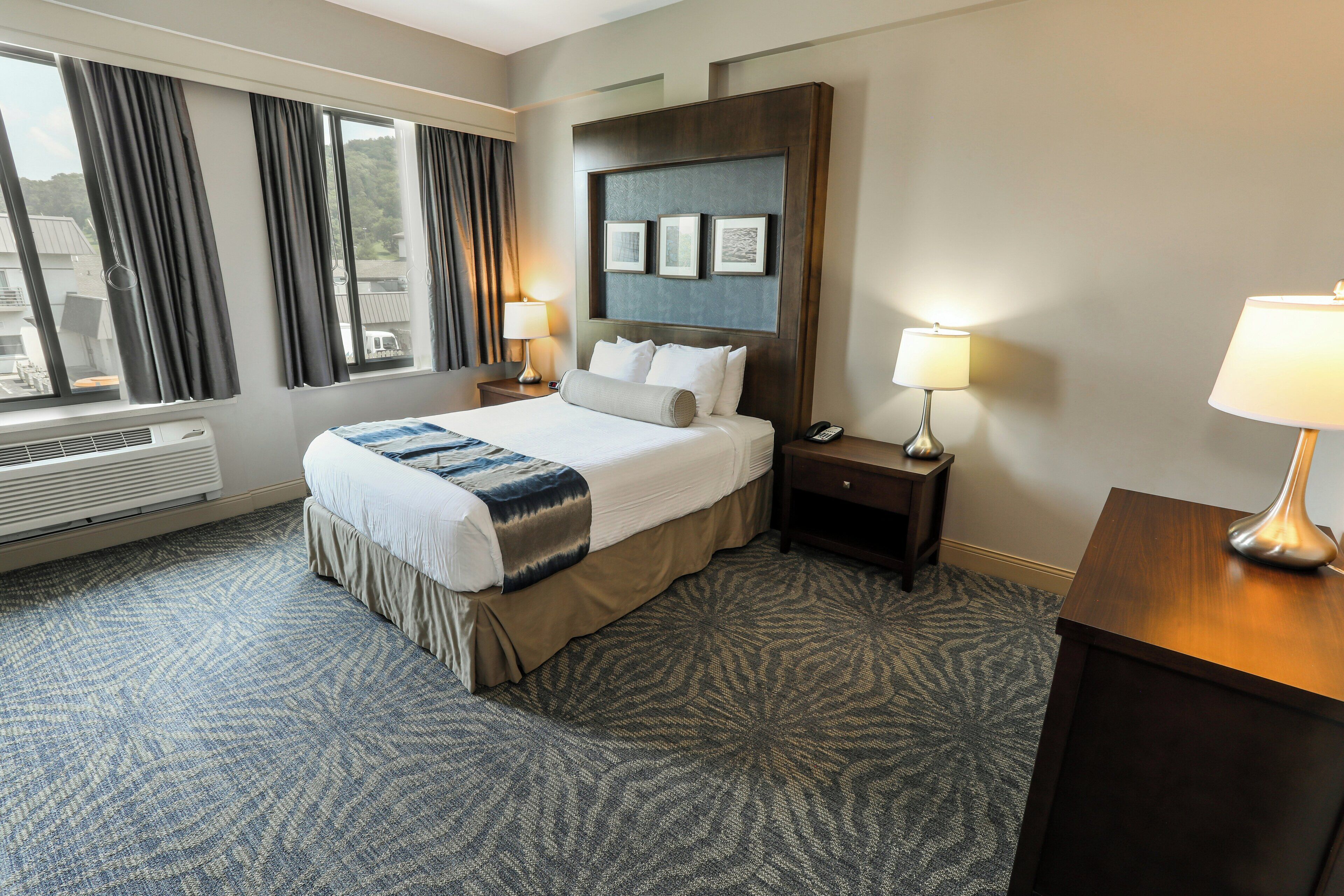 A hotel room with a large bed, two bedside tables with lamps, a large window with curtains, and a wall-mounted picture frame.