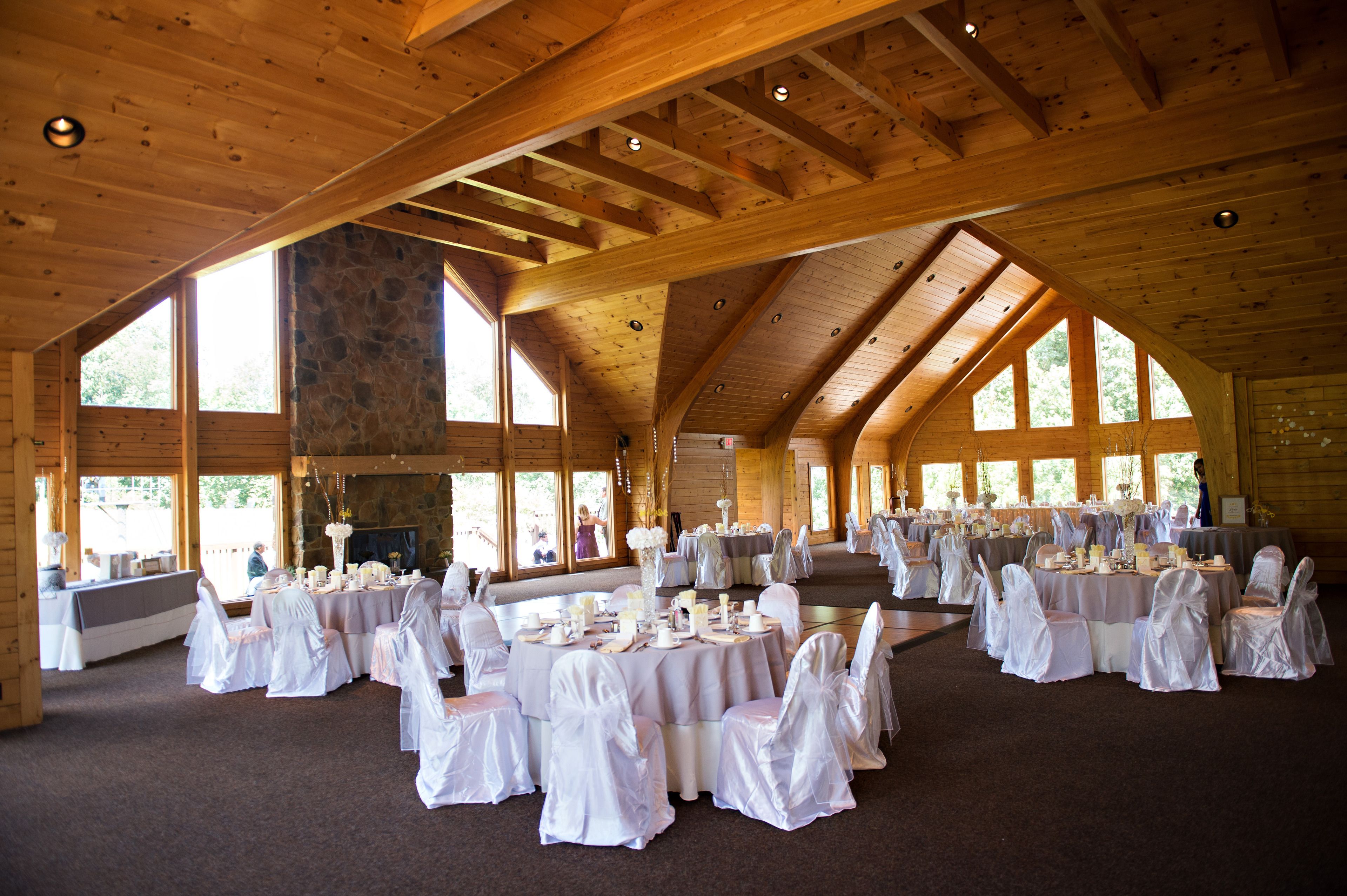 A large hall with high ceilings and wooden beams, furnished with round tables and white chairs, suitable for events.