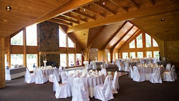 A large hall with high ceilings and wooden beams, furnished with round tables and white chairs, suitable for events.