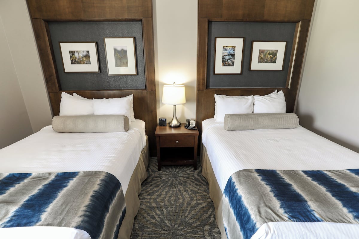 A hotel room with two beds, a nightstand with a lamp, and framed pictures on the walls.
