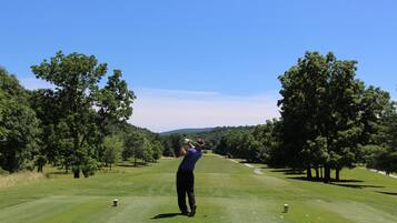 A golf course with a golfer in mid-swing, trees, and a clear sky.