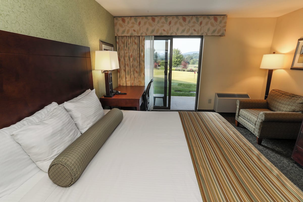 A hotel room with a large bed, a desk, a chair, and a view of a golf course through the sliding glass door.