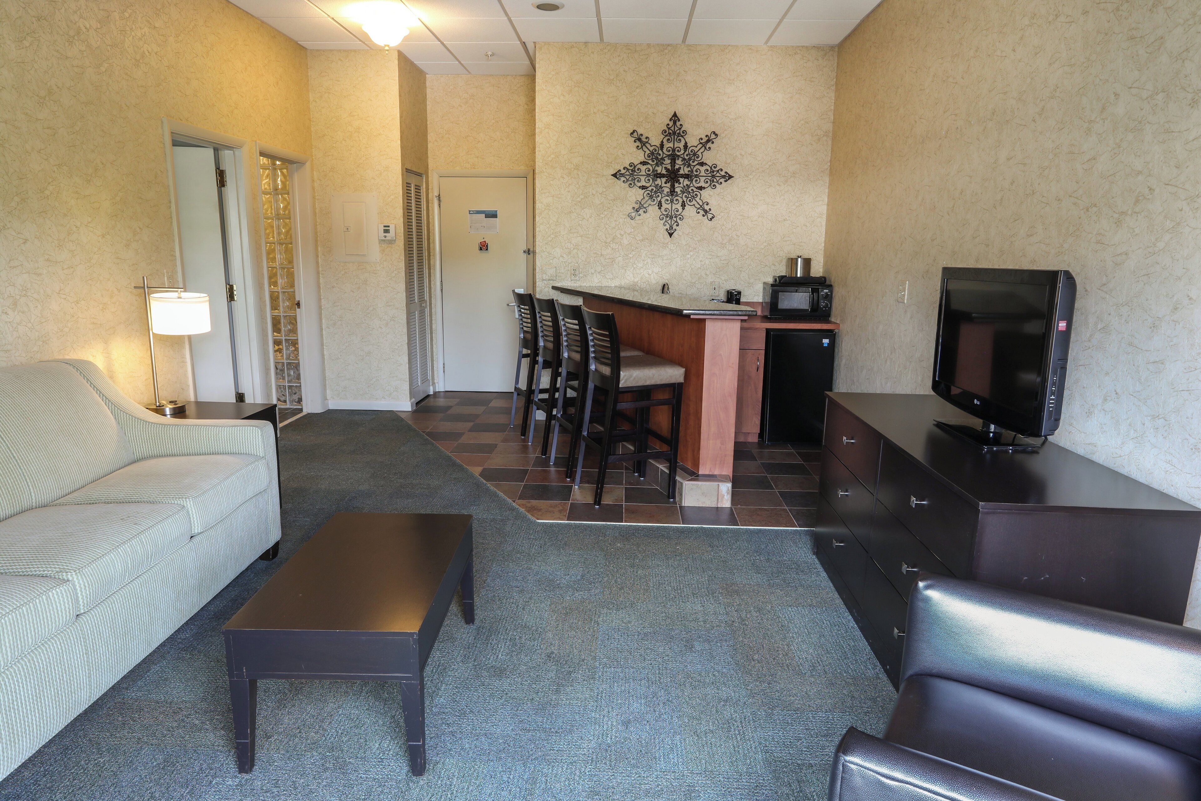 A hotel room with a sofa, a small table, a TV, a kitchenette, and a dining area with chairs.