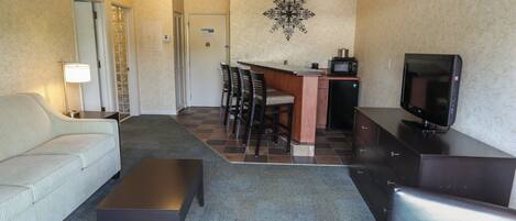 A hotel room with a sofa, a small table, a TV, a kitchenette, and a dining area with chairs.