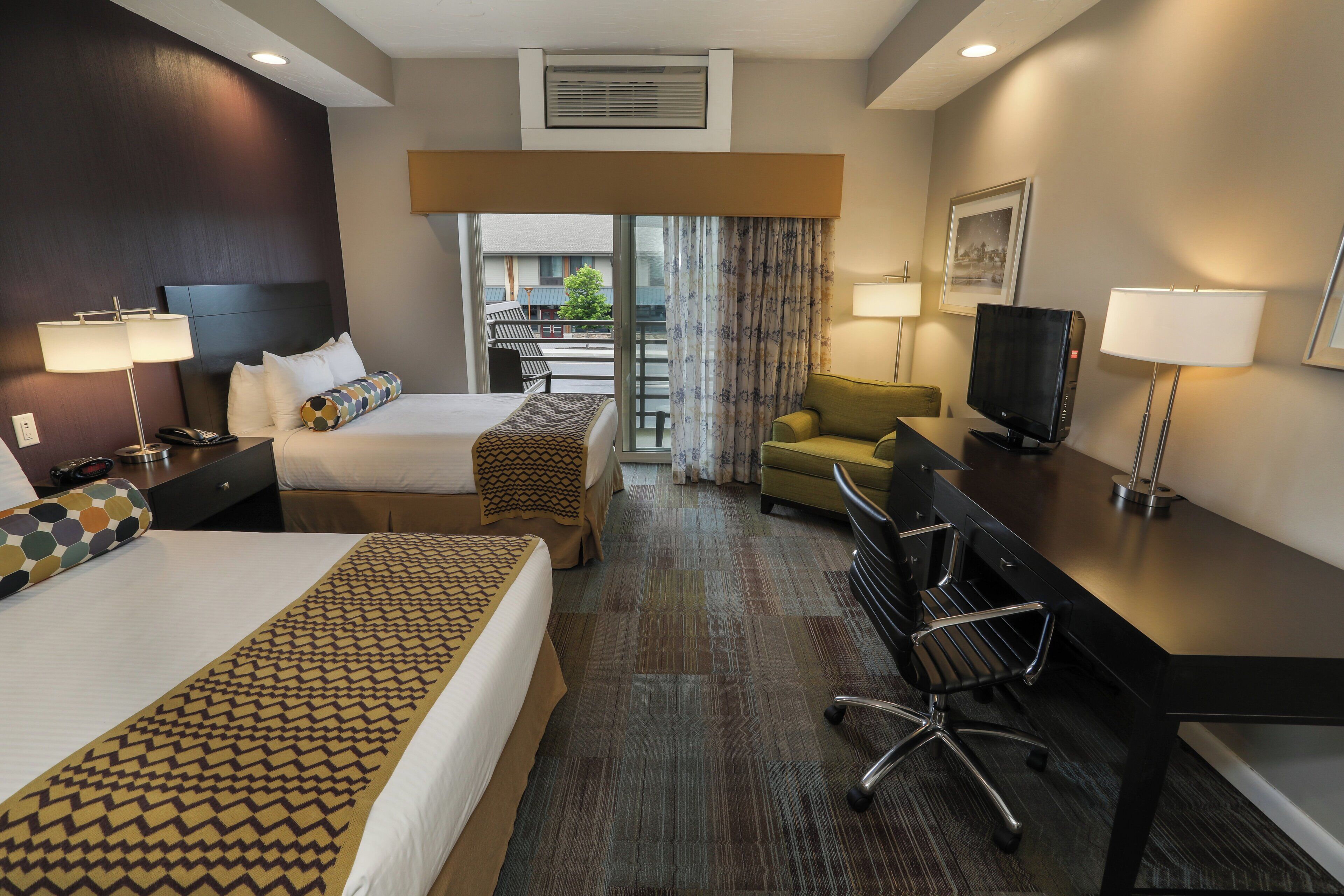 A hotel room with two beds, a desk, a television, and a view of the outside.