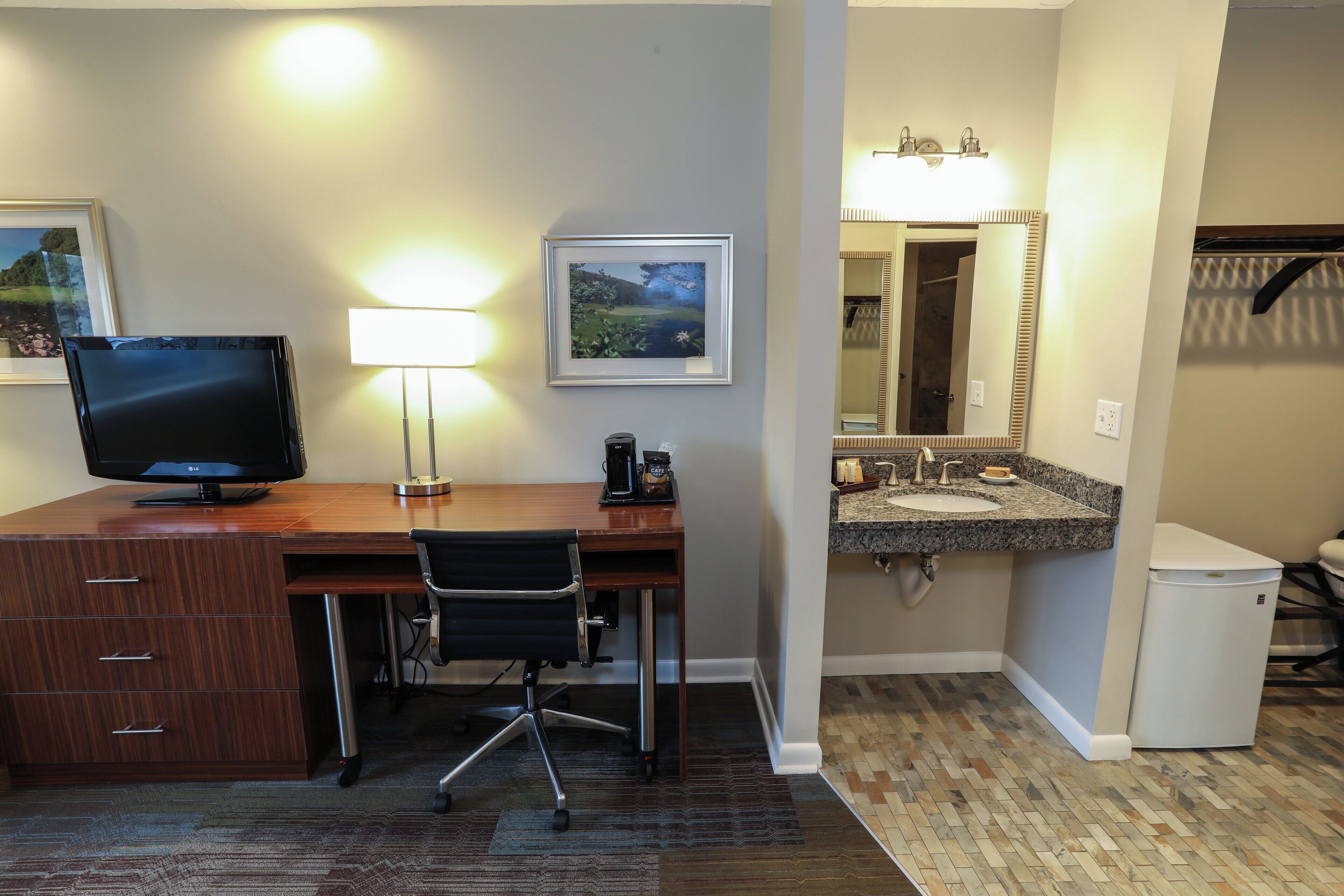 A hotel room with a wooden desk, a computer, a chair, a lamp, a framed picture, a sink, a mirror, and a refrigerator.