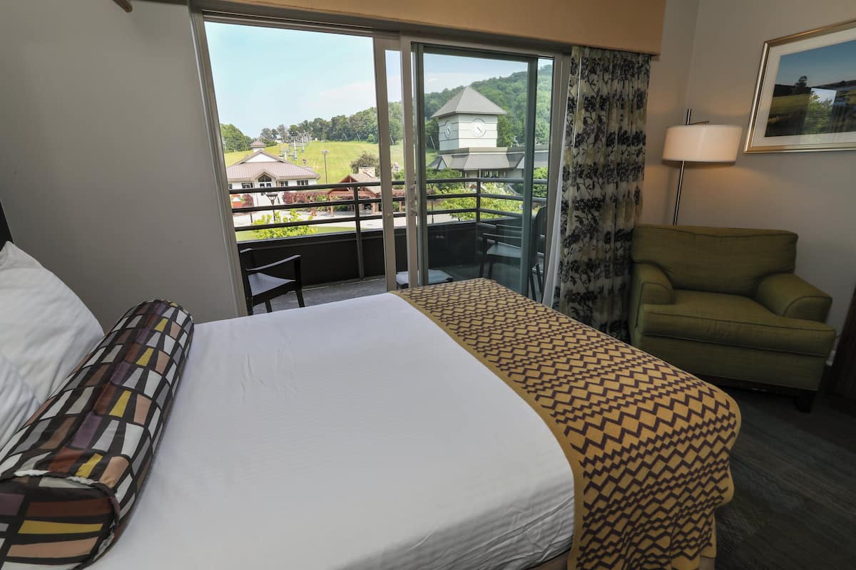 A hotel room with a bed, a green armchair, a table, and a view of a building and greenery outside.