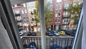 Une vue depuis une fenêtre donnant sur une rue où sont garées des voitures, et sur une rangée d’immeubles en briques rouges, avec des encadrements de fenêtres blancs et des balcons.