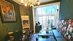 Un espace repas avec une longue table et des bancs, un lustre, une cheminée et une grande fenêtre.