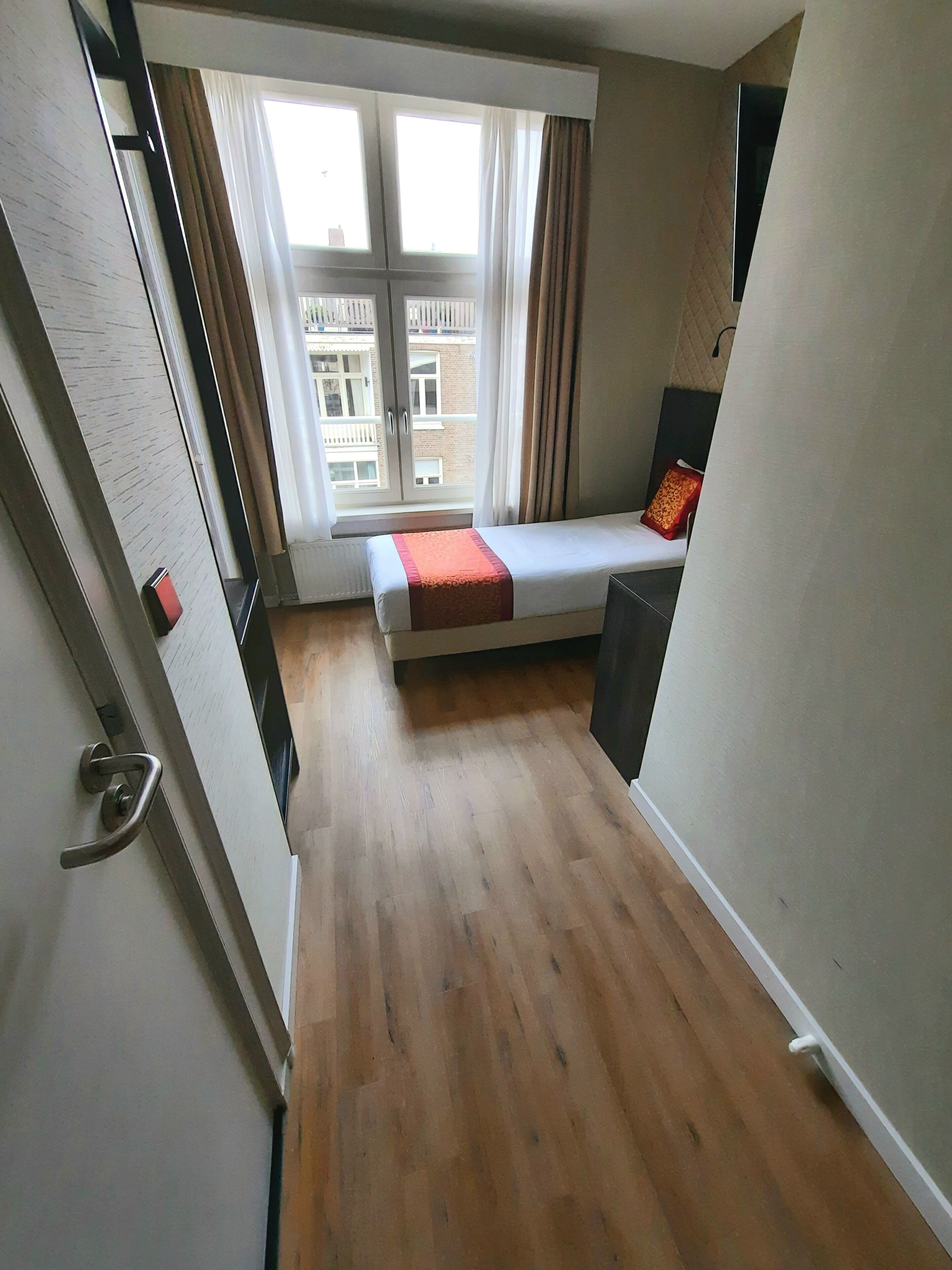 A small, compact room with a bed, a window, and a view of buildings outside.