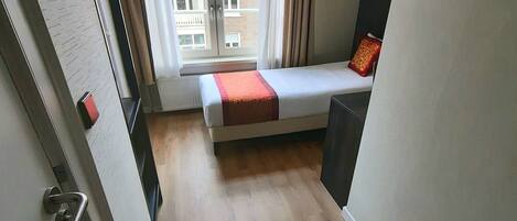 A small, compact room with a bed, a window, and a view of buildings outside.
