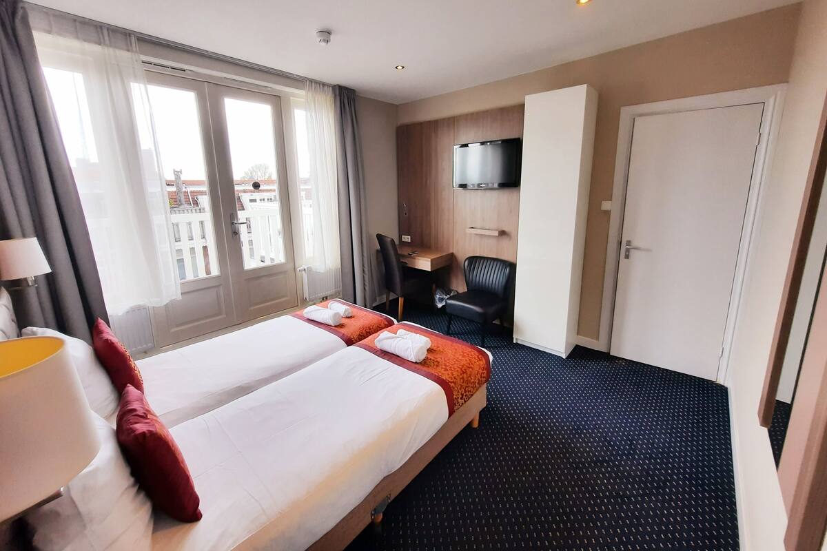 A hotel room with two beds, a desk, a chair, a TV, and a view of the city.