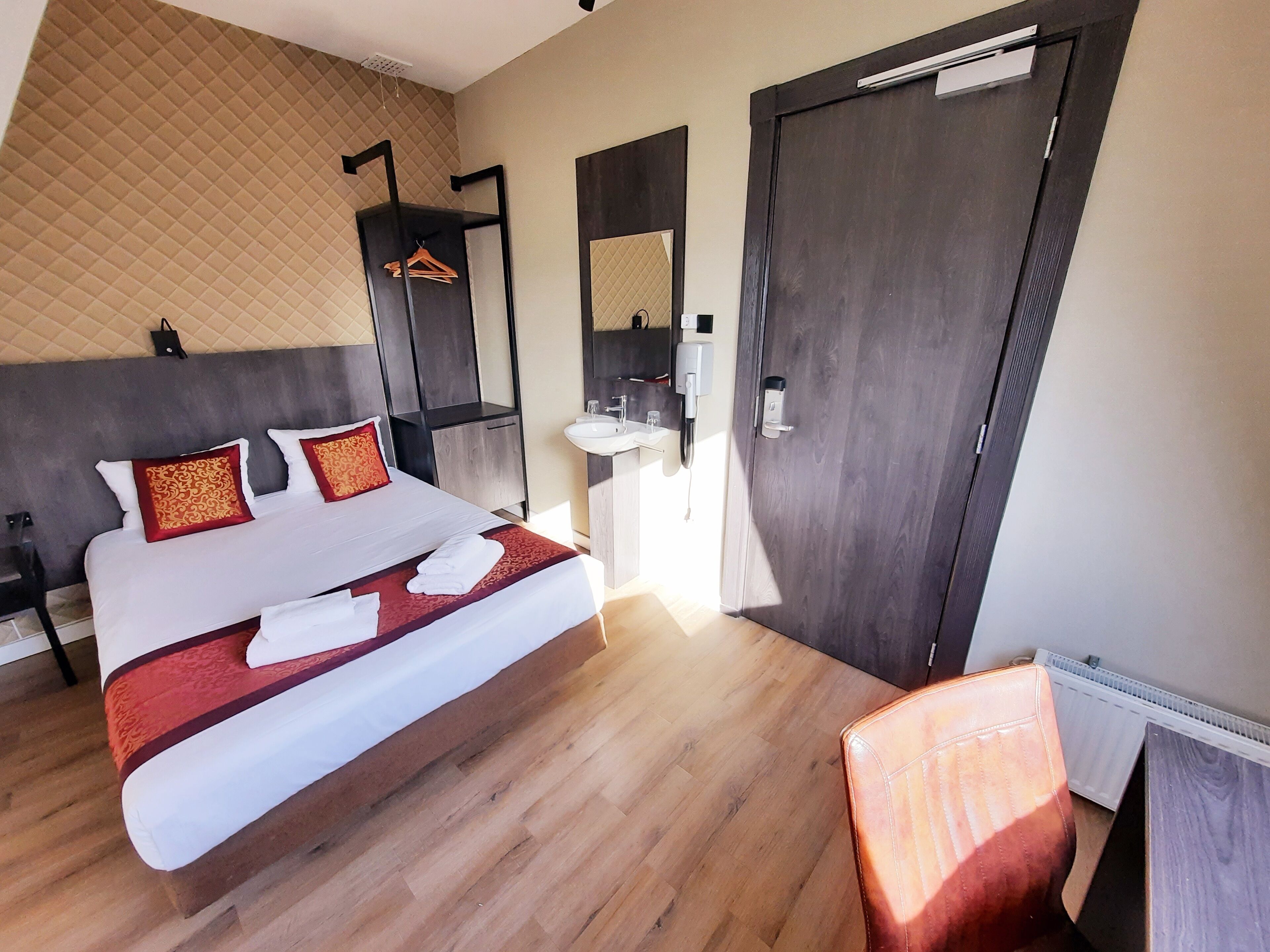 A hotel room with a large bed, a bedside table, a chair, a wardrobe, a mirror, and a bathroom with a sink and toilet.