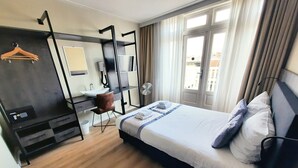 Une chambre d’hôtel comprenant un lit, un bureau, une chaise, un miroir, un porte-vêtements et une fenêtre avec des rideaux.