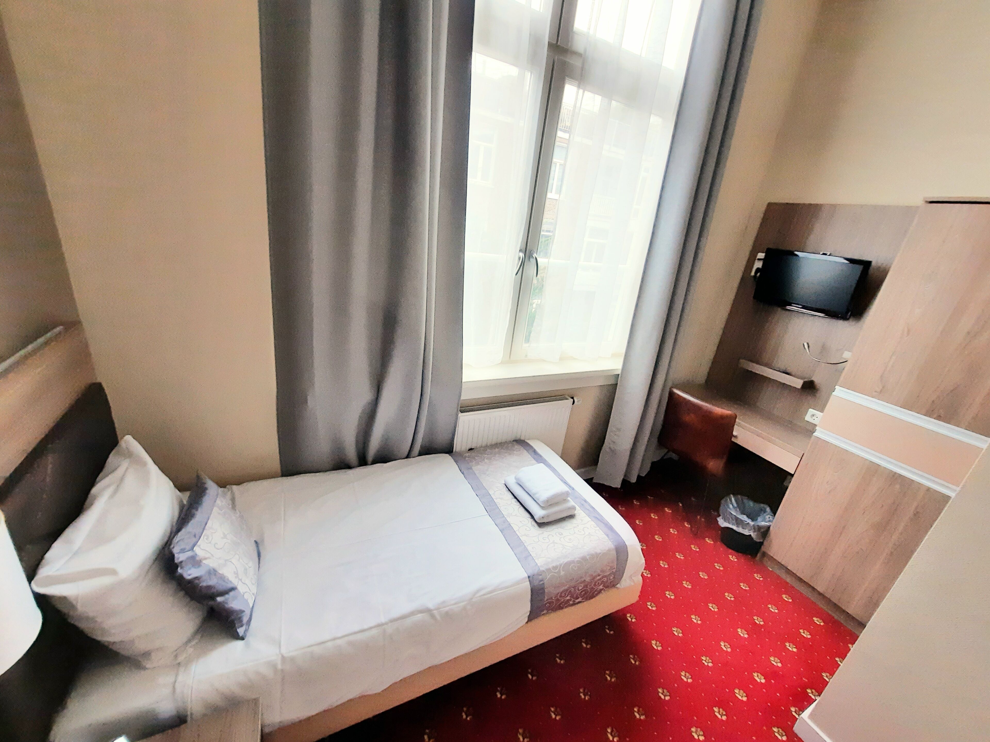 A hotel room with a single bed, a television on a wooden cabinet, and a window with curtains.