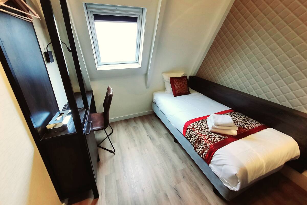 A small, neatly arranged bedroom with a single bed, a desk, a chair, and a window.