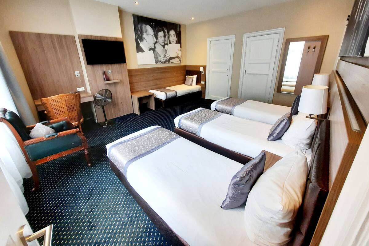 A hotel room with two beds, a sitting area with a chair and a table, a flat-screen TV mounted on the wall, and a large painting above the bed.