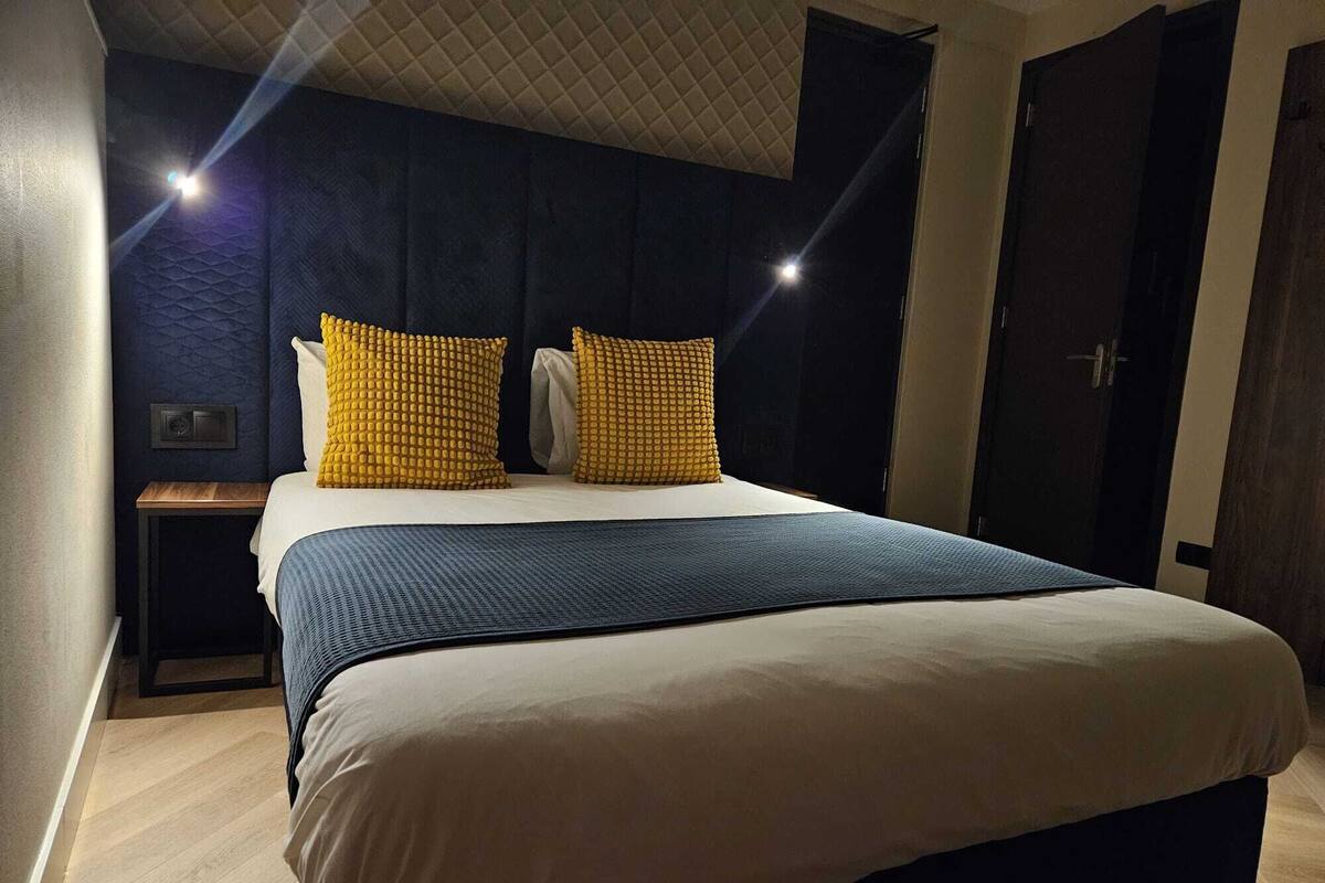A neatly made bed with a blue and white duvet, two yellow textured pillows, a bedside table with a lamp, and a dark wooden wardrobe.