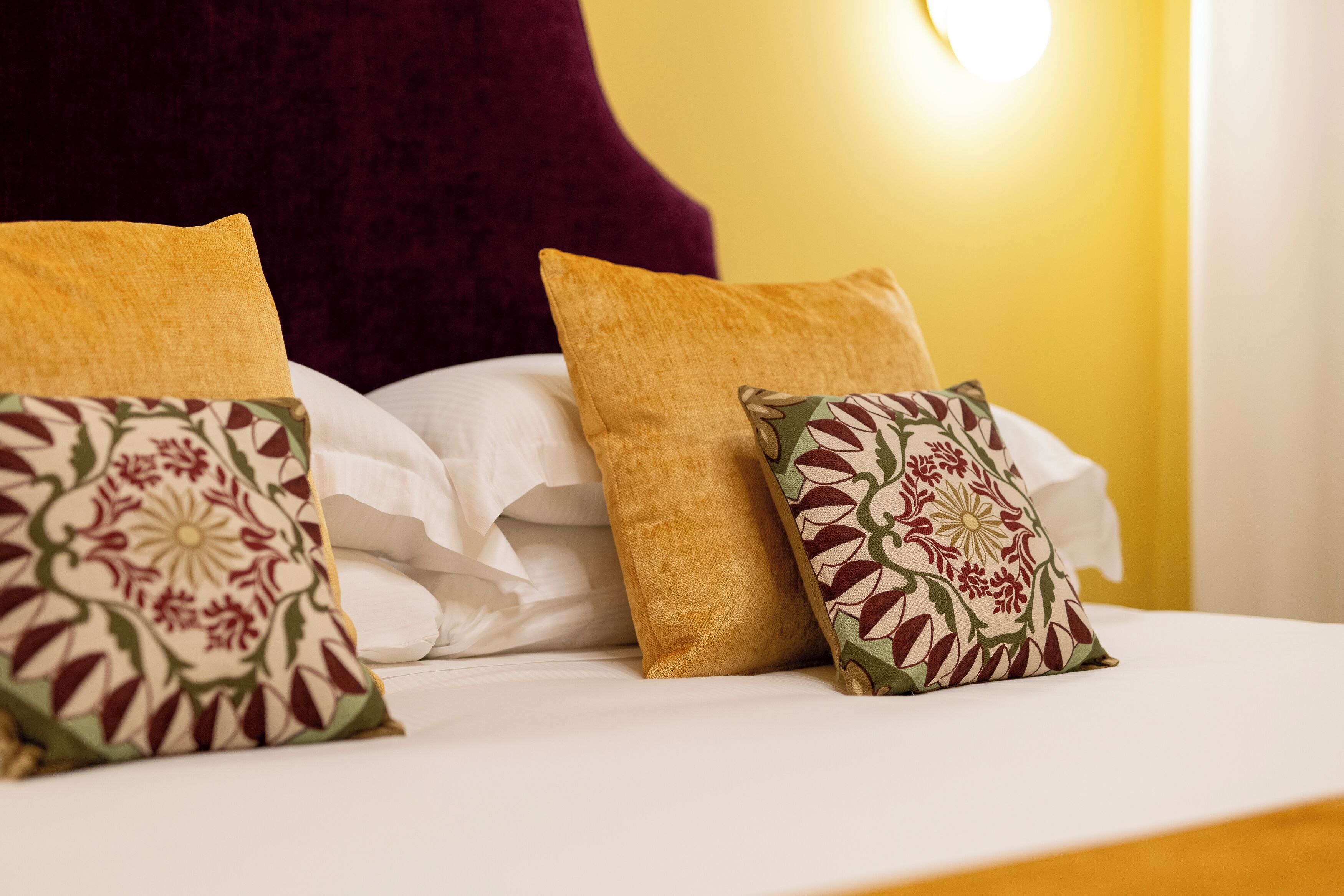 Una cama con sábanas blancas y almohadones decorativos, un cabecero rojo oscuro y una pared amarilla con una lámpara.